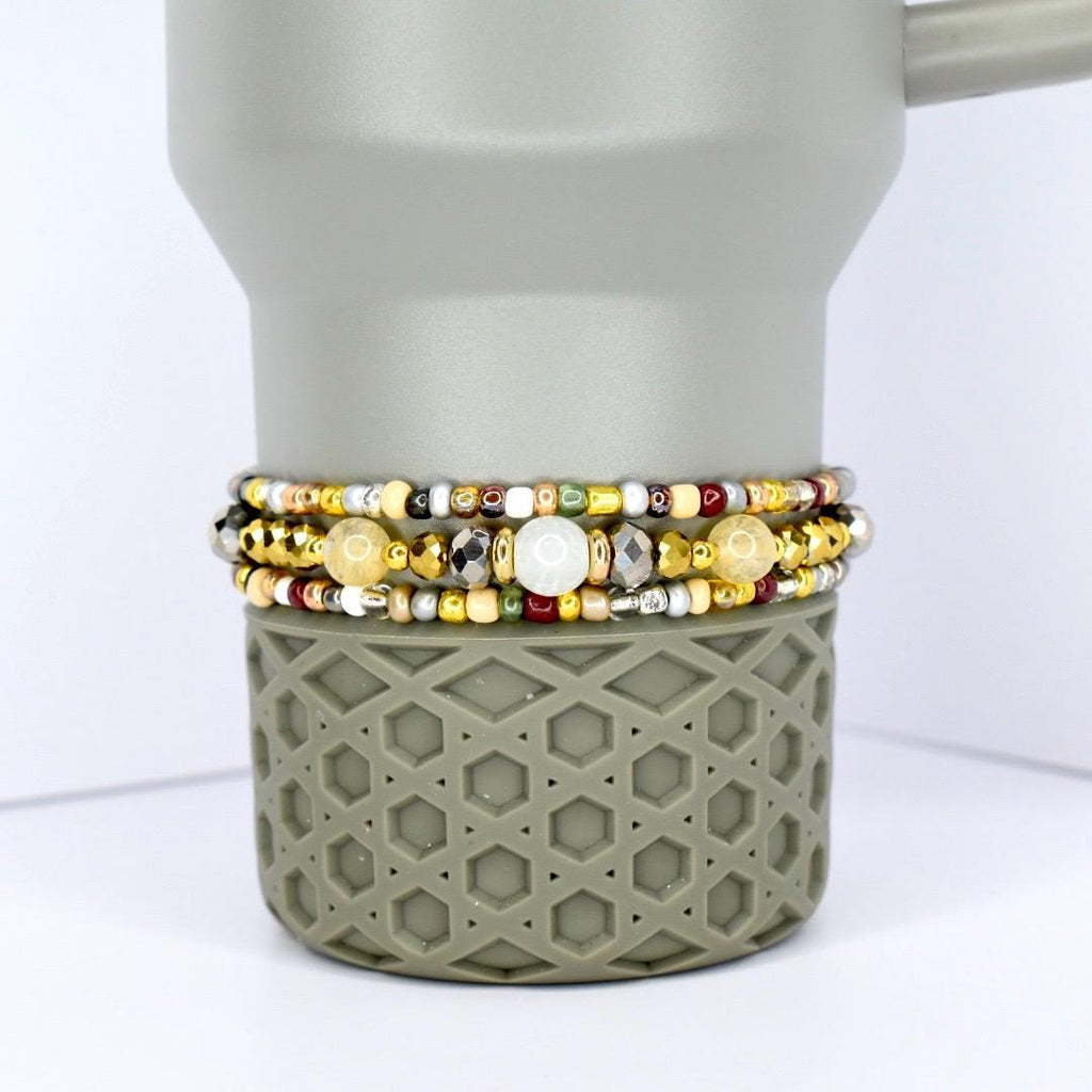 Three beaded bracelets with gold, silver, and colorful beads are stacked on a gray tumbler. The bracelets feature a variety of bead sizes and colors, including white, yellow, and red. The tumbler has a geometric pattern.