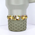 Three beaded bracelets with gold, silver, and colorful beads are stacked on a gray tumbler. The bracelets feature a variety of bead sizes and colors, including white, yellow, and red. The tumbler has a geometric pattern.