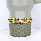 Three beaded bracelets with gold, silver, and colorful beads are stacked on a gray tumbler. The bracelets feature a variety of bead sizes and colors, including white, yellow, and red. The tumbler has a geometric pattern.