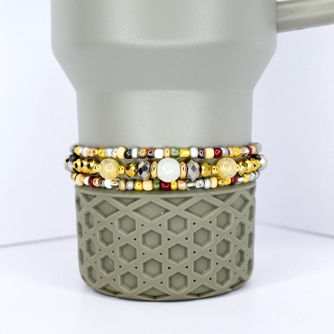 Three beaded bracelets with gold, silver, and colorful beads are stacked on a gray tumbler. The bracelets feature a variety of bead sizes and colors, including white, yellow, and red. The tumbler has a geometric pattern.