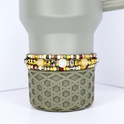 Three beaded bracelets with gold, silver, and colorful beads are stacked on a gray tumbler. The bracelets feature a variety of bead sizes and colors, including white, yellow, and red. The tumbler has a geometric pattern.