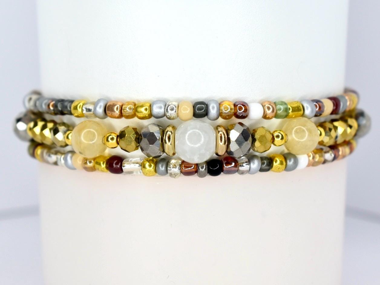Three beaded bracelets with a variety of colors and textures. The bracelets feature gold, silver, and bronze beads, as well as larger, faceted beads and round, white beads. The bracelets are stacked together.