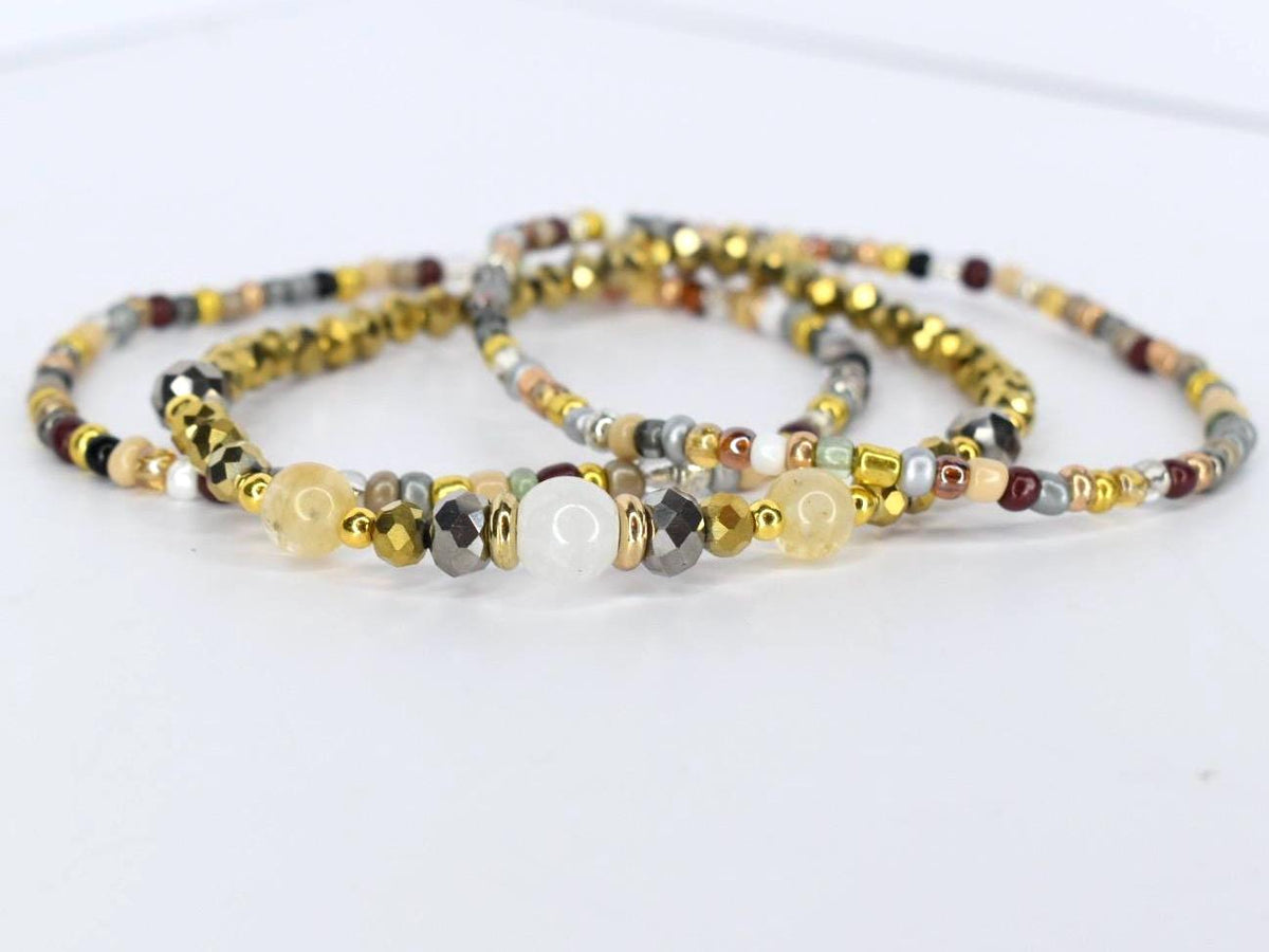 Three beaded bracelets in gold, silver, and neutral tones. The bracelets feature a variety of beads, including faceted metallic beads, small round beads, and larger, smooth beads in yellow and white.