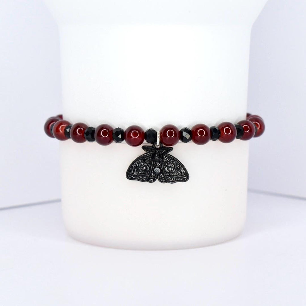 A beaded bracelet with a black moth charm. The bracelet features dark red, round beads interspersed with smaller, black beads. The moth charm is black and has intricate wing detailing, hanging from the center of the bracelet.