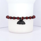 A beaded bracelet with a black moth charm. The bracelet features dark red, round beads interspersed with smaller, black beads. The moth charm is black and has intricate wing detailing, hanging from the center of the bracelet.