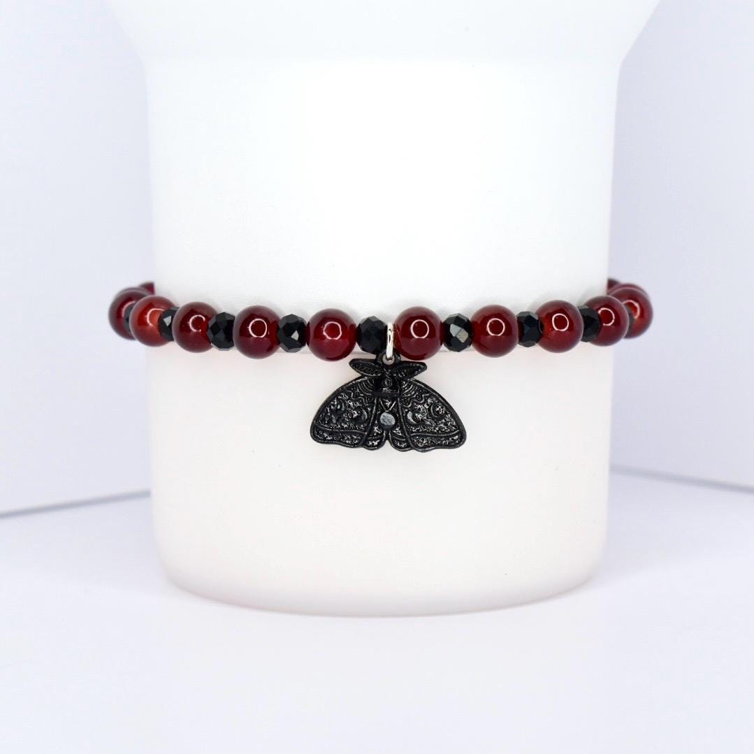 A beaded bracelet with a black moth charm. The bracelet features dark red, round beads interspersed with smaller, black beads. The moth charm is black and has intricate wing detailing, hanging from the center of the bracelet.