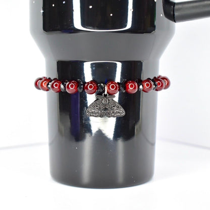 A dark red beaded bracelet encircles a black tumbler. The bracelet features small, round, dark red beads interspersed with black beads. A silver-colored moth charm hangs from the bracelet. The tumbler is black and has a handle.