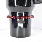 A dark red beaded bracelet encircles a black tumbler. The bracelet features small, round, dark red beads interspersed with black beads. A silver-colored moth charm hangs from the bracelet. The tumbler is black and has a handle.