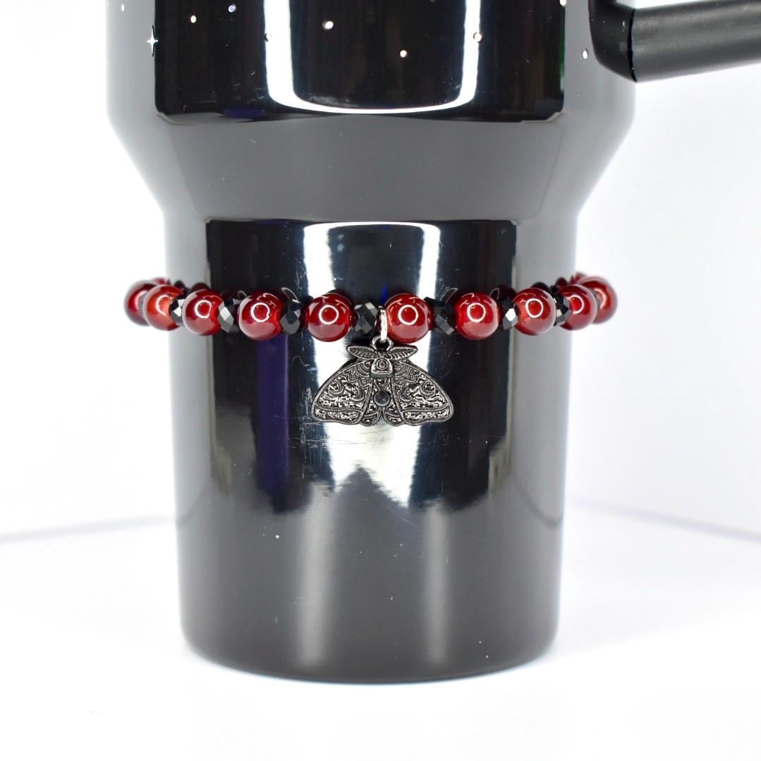 A dark red beaded bracelet encircles a black tumbler. The bracelet features small, round, dark red beads interspersed with black beads. A silver-colored moth charm hangs from the bracelet. The tumbler is black and has a handle.