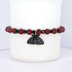 A beaded bracelet featuring dark red, round beads interspersed with black, faceted beads. A black moth charm hangs from the center. The bracelet is displayed against a white background.