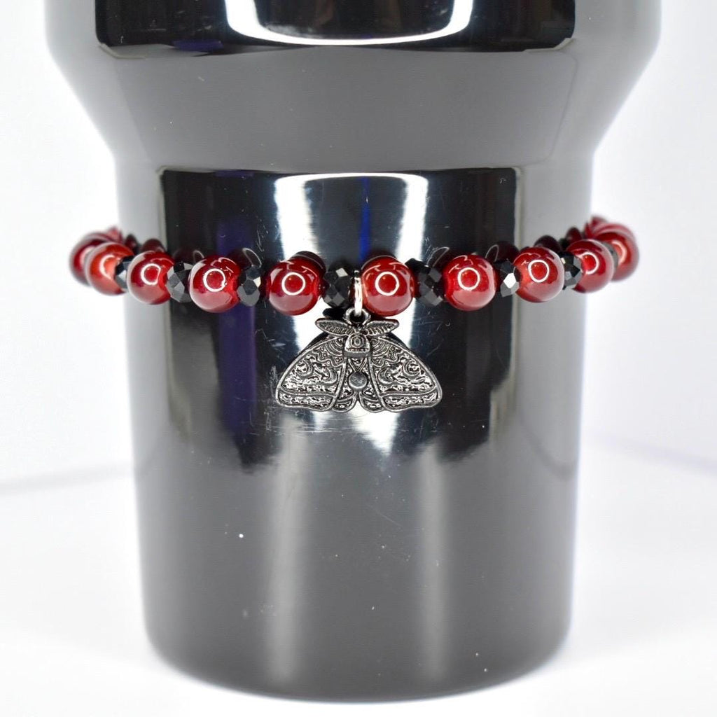 A dark red beaded bracelet with a silver moth pendant. The bracelet features round, glossy red beads interspersed with smaller black beads. The moth pendant is detailed with intricate wing and body patterns, adding a gothic or alternative aesthetic to the jewelry.