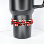 Three beaded bracelets wrapped around a black tumbler. The bracelets feature red, black, and clear beads with silver accents. The beads are arranged in various patterns, creating a layered effect. The tumbler has a sleek, matte finish and a handle.