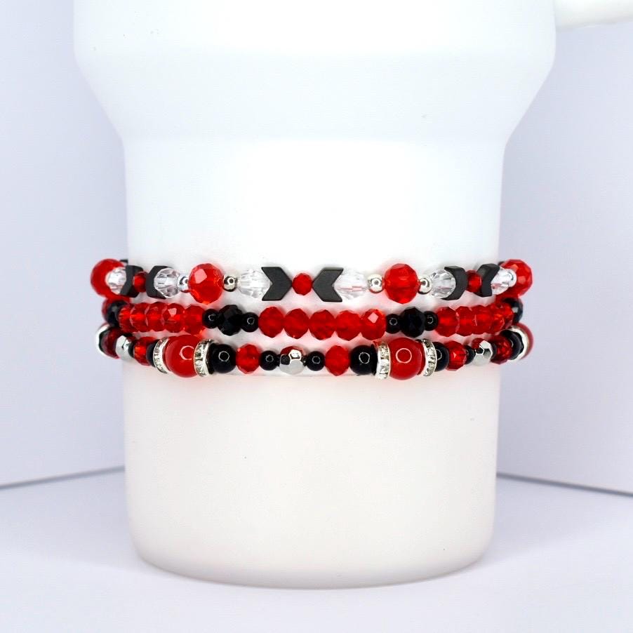 Three beaded bracelets in red, black, and silver. The top bracelet features red and black beads with silver accents and black arrow-shaped beads. The middle bracelet has red and black beads with silver accents. The bottom bracelet is primarily red with black and silver beads.