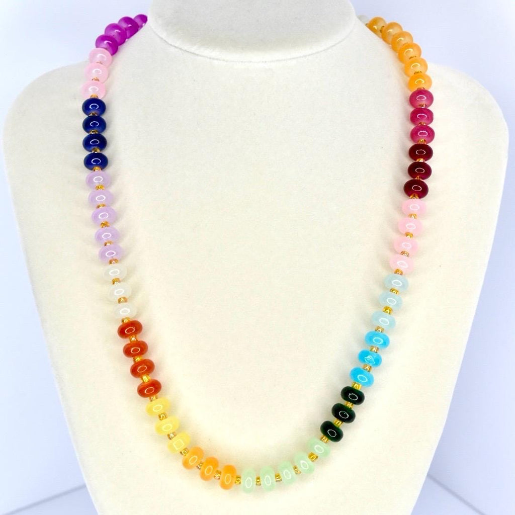 A colorful beaded necklace displayed on a white mannequin. The necklace features a gradient of colors, including purple, pink, blue, green, yellow, orange, and red. The beads are round and strung together with gold-colored string.