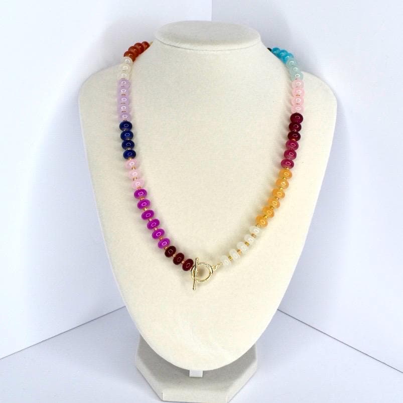 A colorful beaded necklace displayed on a white bust. The necklace features a gradient of colors, including red, orange, yellow, green, blue, and purple beads. It has a gold-colored toggle clasp.