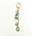 Gold-tone keychain with a lobster clasp and a chain holding several charms. Charms include a seashell, a blue leaf, a heart with pearls, a large pearl, a fan-shaped charm, and an oval charm with a beach scene.