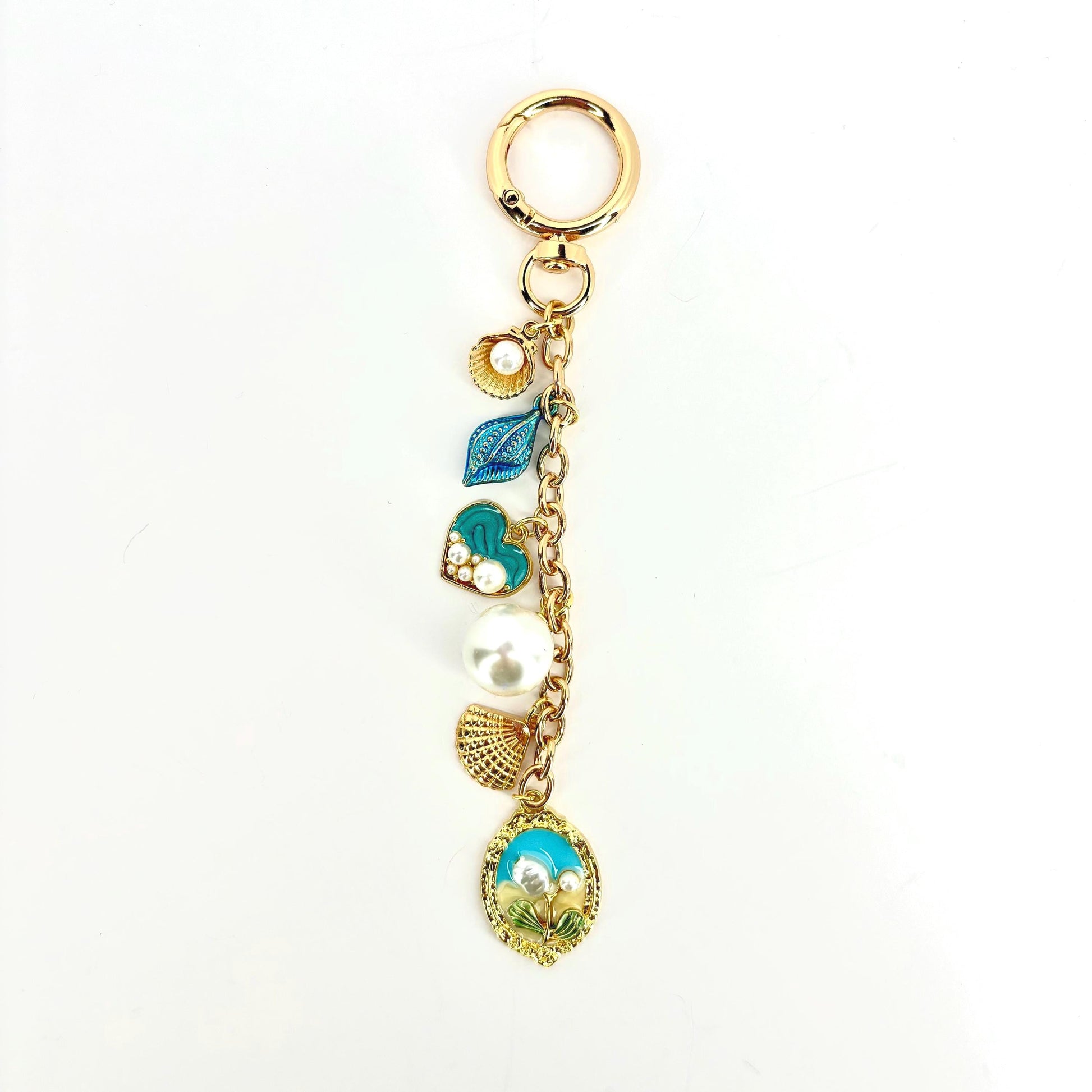 Gold-tone keychain with a lobster clasp and a chain holding several charms. Charms include a seashell, a blue leaf, a heart with pearls, a large pearl, a fan-shaped charm, and an oval charm with a beach scene.