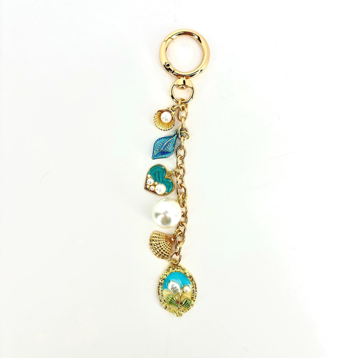 Gold-tone keychain with a lobster clasp and a chain holding several charms. Charms include a seashell, a blue leaf, a heart with pearls, a large pearl, a fan-shaped charm, and an oval charm with a beach scene.