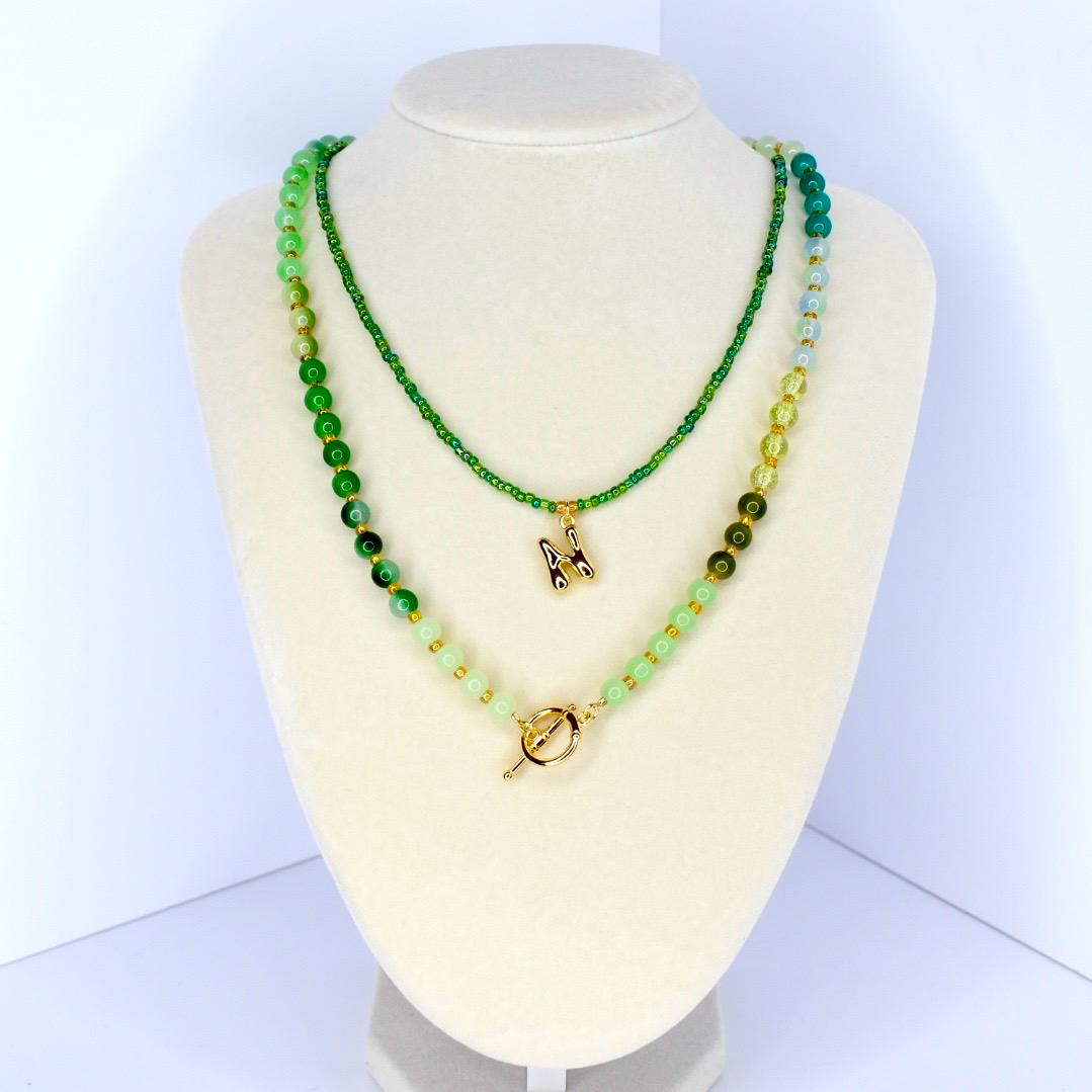Two-strand necklace set displayed on a white mannequin. The necklaces feature green beads and a gold-colored initial pendant. The lower necklace has a gold toggle clasp. The beads vary in size and shade, creating a layered effect.