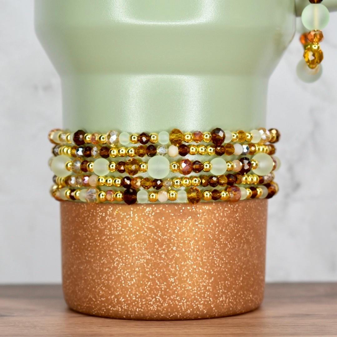 A stack of beaded bracelets in shades of brown, gold, and cream, with a mix of small and large beads. The bracelets are displayed on a copper-colored, glittery cylindrical base, with a light green background.