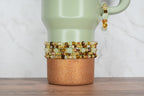 A light green travel mug with a copper glitter base. Several beaded bracelets in shades of gold, brown, and green are stacked around the mug. A matching bracelet hangs from the handle.