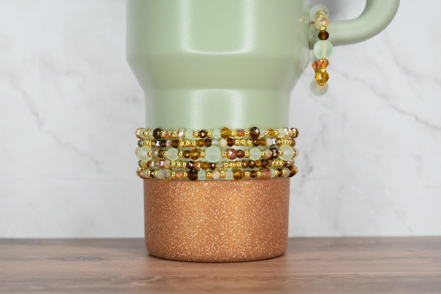 A light green travel mug with a copper glitter base. Several beaded bracelets in shades of gold, brown, and green are stacked around the mug. A matching bracelet hangs from the handle.