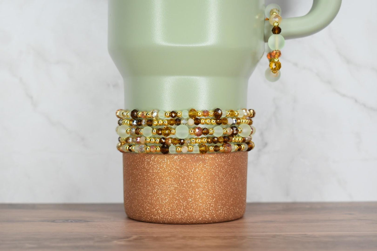 A light green travel mug with a copper glitter base. Several beaded bracelets in shades of gold, brown, and green are stacked around the mug. A matching bracelet hangs from the handle.