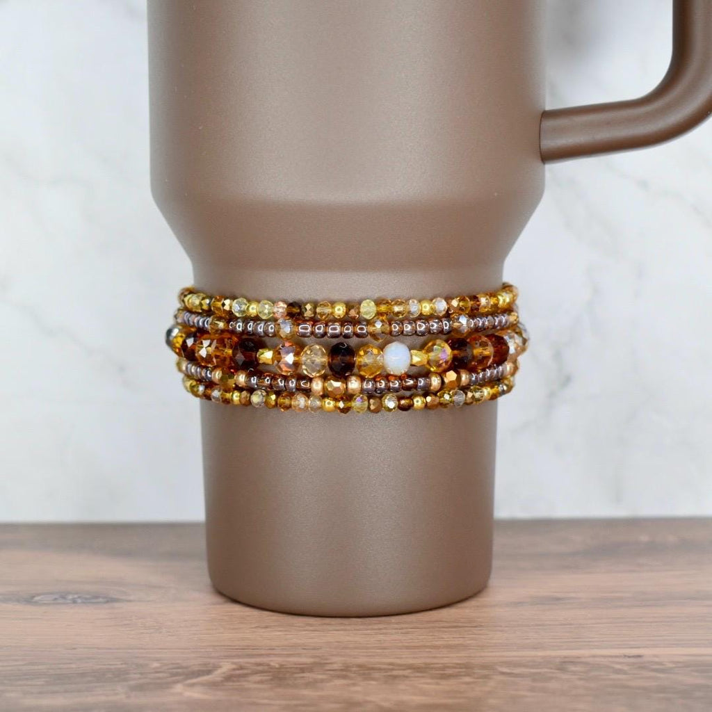 A brown travel mug with a beaded wrap. The wrap features multiple strands of amber, gold, and brown beads. The mug has a handle and sits on a wooden surface.