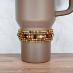 A brown travel mug with a beaded wrap. The wrap features multiple strands of amber, gold, and brown beads. The mug has a handle and sits on a wooden surface.