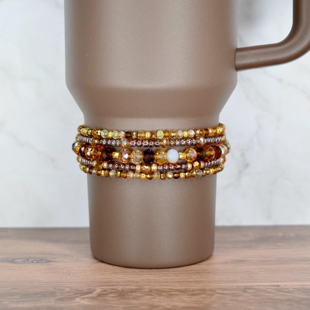 A brown travel mug with a beaded wrap. The wrap features multiple strands of amber, gold, and brown beads. The mug has a handle and sits on a wooden surface.