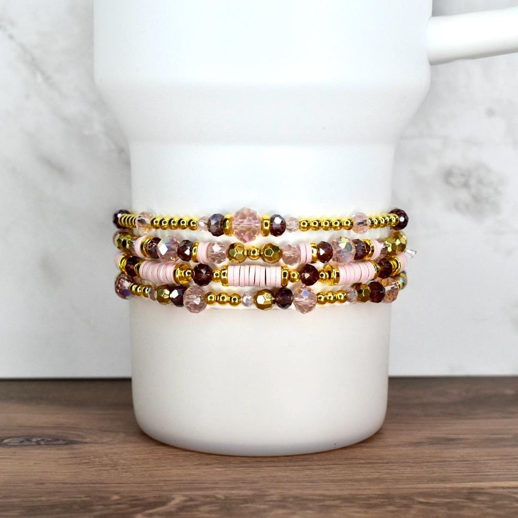 Four beaded bracelets in shades of pink, gold, and purple are stacked on a white cylindrical object. The bracelets feature a variety of beads, including small gold beads, faceted crystals, and larger pink beads. The bracelets are displayed on a white background.
