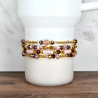 Four beaded bracelets in shades of pink, gold, and purple are stacked on a white cylindrical object. The bracelets feature a variety of beads, including small gold beads, faceted crystals, and larger pink beads. The bracelets are displayed on a white background.