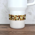 A white tumbler with a beaded wrap. The wrap features multiple strands of small, faceted beads in shades of brown, gold, and cream. The beads are strung together with gold-colored wire, creating a decorative accessory.