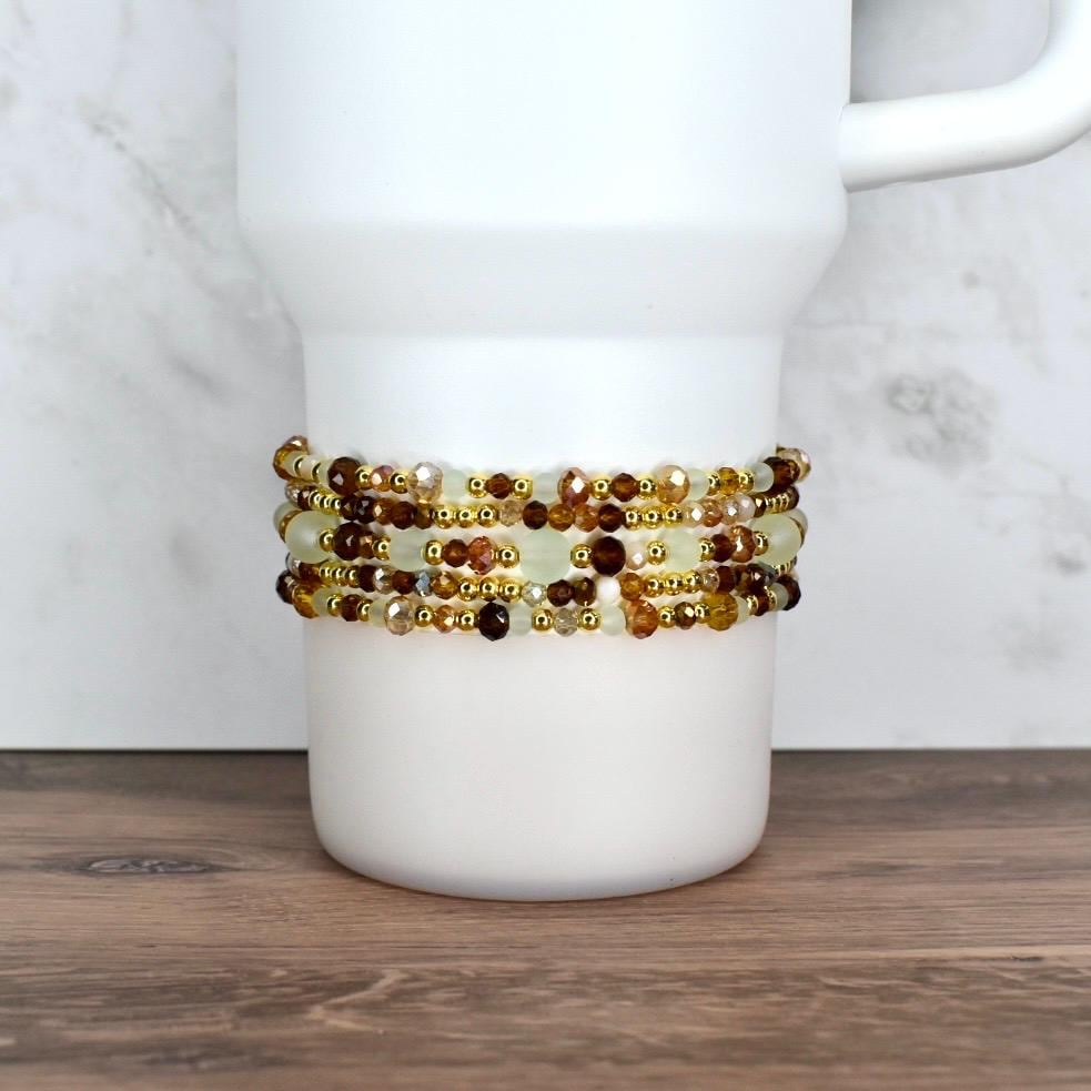 A white tumbler with a beaded wrap. The wrap features multiple strands of small, faceted beads in shades of brown, gold, and cream. The beads are strung together with gold-colored wire, creating a decorative accessory.