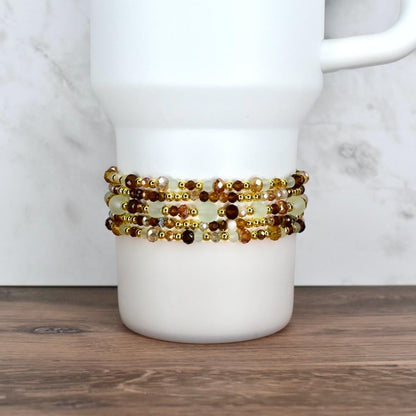 A white tumbler with a beaded wrap. The wrap features multiple strands of small, faceted beads in shades of brown, gold, and cream. The beads are strung together with gold-colored wire, creating a decorative accessory.