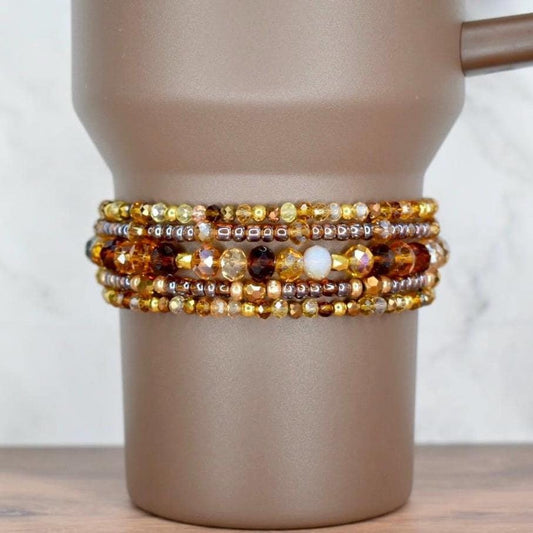 A set of five beaded bracelets on the base of a brown Stanley tumbler. The top and bottom bracelets have a mix of gold, brown, and amber seed beads. The two bracelets surrounding the middle bracelet feature transparent brown seed beads and sparkly glass beads in brown, bronze, and amber. The middle bracelet has mid-sized glass beads in different shades of brown and cream.