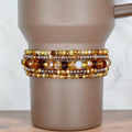 A set of five beaded bracelets on the base of a brown Stanley tumbler. The top and bottom bracelets have a mix of gold, brown, and amber seed beads. The two bracelets surrounding the middle bracelet feature transparent brown seed beads and sparkly glass beads in brown, bronze, and amber. The middle bracelet has mid-sized glass beads in different shades of brown and cream.