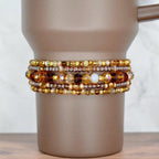 A set of five beaded bracelets on the base of a brown Stanley tumbler. The top and bottom bracelets have a mix of gold, brown, and amber seed beads. The two bracelets surrounding the middle bracelet feature transparent brown seed beads and sparkly glass beads in brown, bronze, and amber. The middle bracelet has mid-sized glass beads in different shades of brown and cream.