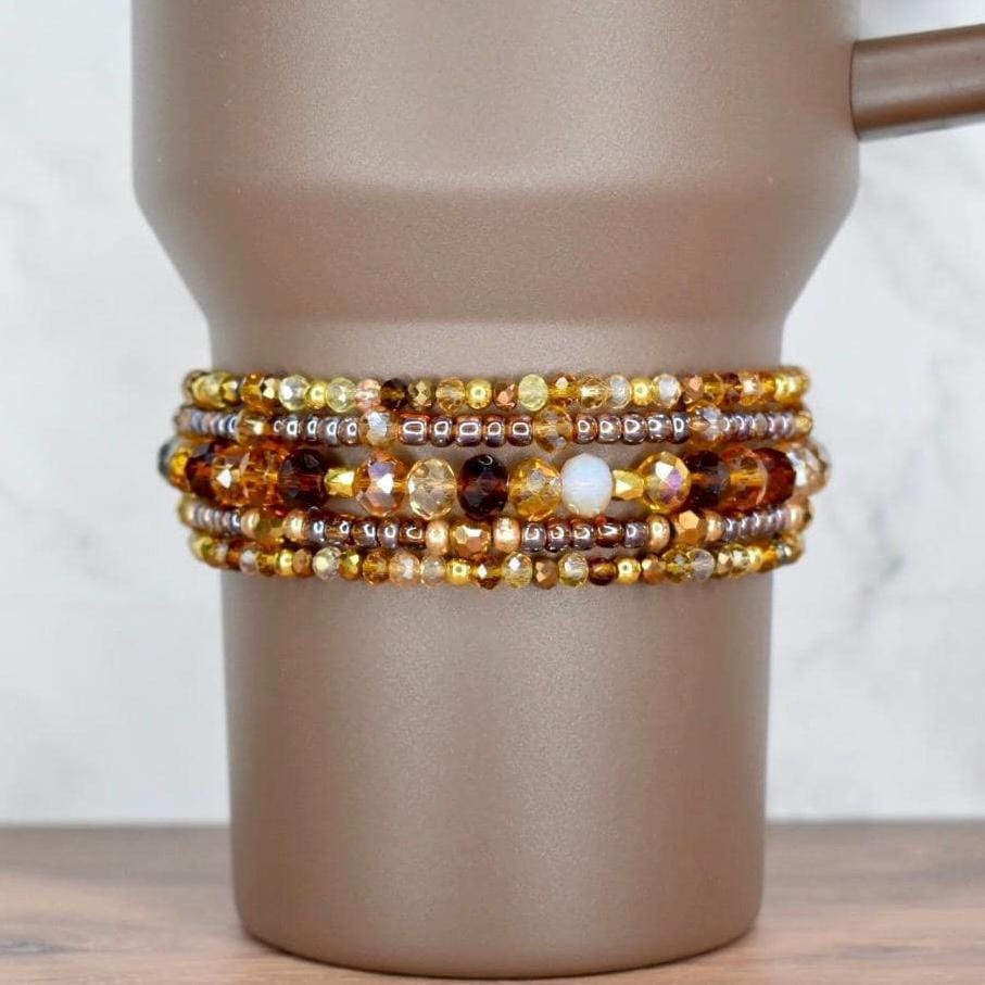 A set of five beaded bracelets on the base of a brown Stanley tumbler. The top and bottom bracelets have a mix of gold, brown, and amber seed beads. The two bracelets surrounding the middle bracelet feature transparent brown seed beads and sparkly glass beads in brown, bronze, and amber. The middle bracelet has mid-sized glass beads in different shades of brown and cream.