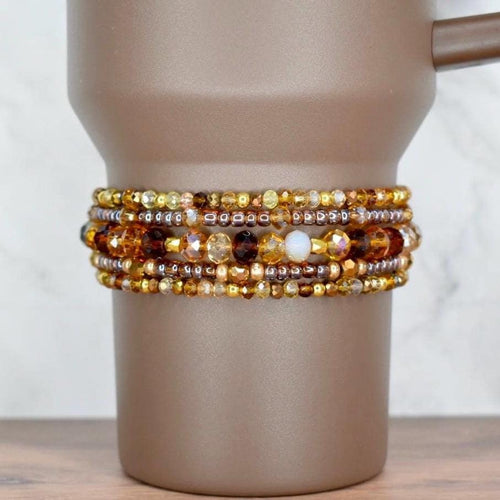 A set of five beaded bracelets on the base of a brown Stanley tumbler. The top and bottom bracelets have a mix of gold, brown, and amber seed beads. The two bracelets surrounding the middle bracelet feature transparent brown seed beads and sparkly glass beads in brown, bronze, and amber. The middle bracelet has mid-sized glass beads in different shades of brown and cream.