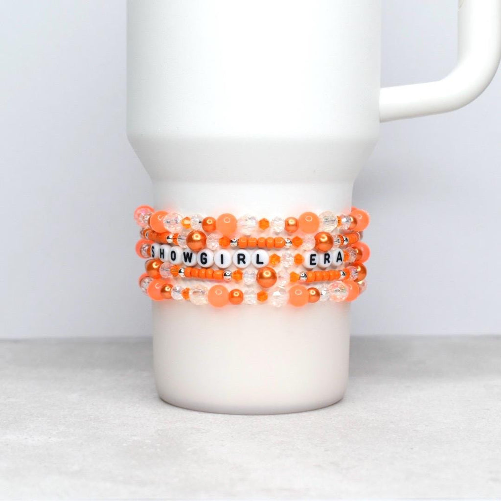 Five orange-toned beaded bracelets featuring a mix of glossy pearls, translucent crystals, and seed beads wrap around a white tumbler. The phrase “SHOWGIRL ERA” is spelled in black lettering on white beads, set against a soft orange bokeh backdrop.