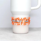 Five orange-toned beaded bracelets featuring a mix of glossy pearls, translucent crystals, and seed beads wrap around a white tumbler. The phrase “SHOWGIRL ERA” is spelled in black lettering on white beads, set against a soft orange bokeh backdrop.