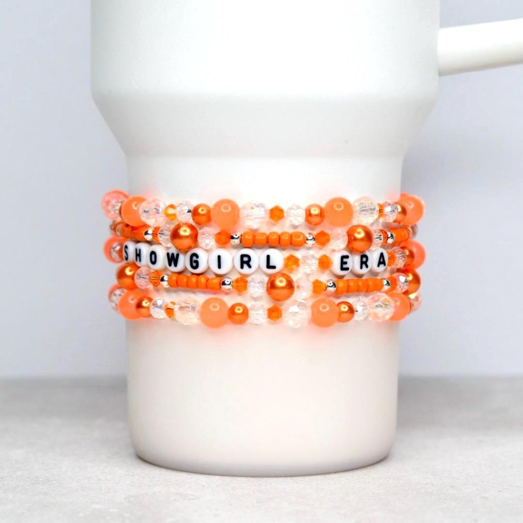 A white tumbler adorned with a stack of orange, peach, and clear beaded bracelets featuring white letter beads that spell “SHOWGIRL ERA.” The mix of pearl, faceted, and seed beads creates a vibrant, sparkly look against a blurred orange bokeh background.