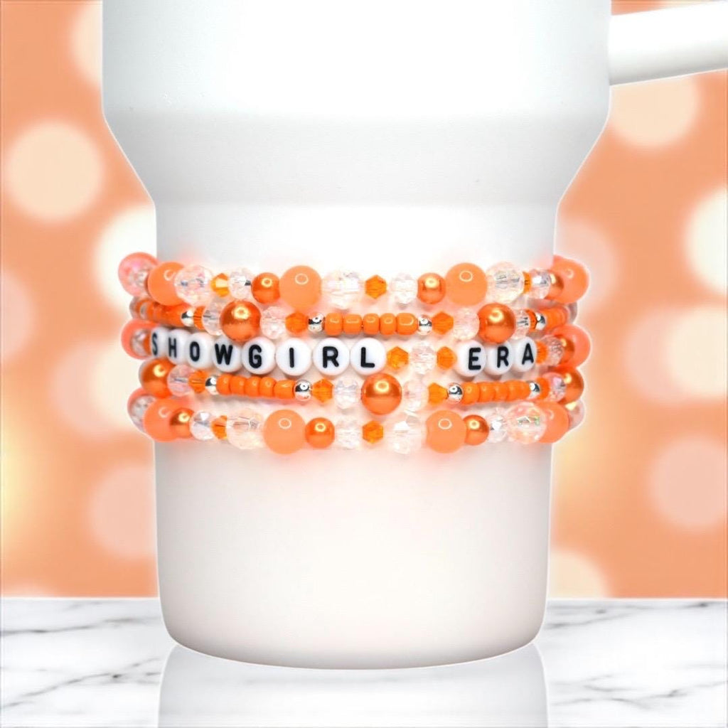 A white Stanley wrapped with a beaded bracelet stack in bright orange, peach, and clear tones. The stack features round pearl, faceted crystal, and opaque seed beads, with white alphabet beads spelling “SHOWGIRL ERA” in black letters across the center. The bracelet glimmers under soft lighting against a warm orange bokeh background, giving a playful, glamorous, and energetic vibe.
