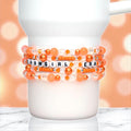 A white Stanley wrapped with a beaded bracelet stack in bright orange, peach, and clear tones. The stack features round pearl, faceted crystal, and opaque seed beads, with white alphabet beads spelling “SHOWGIRL ERA” in black letters across the center. The bracelet glimmers under soft lighting against a warm orange bokeh background, giving a playful, glamorous, and energetic vibe.