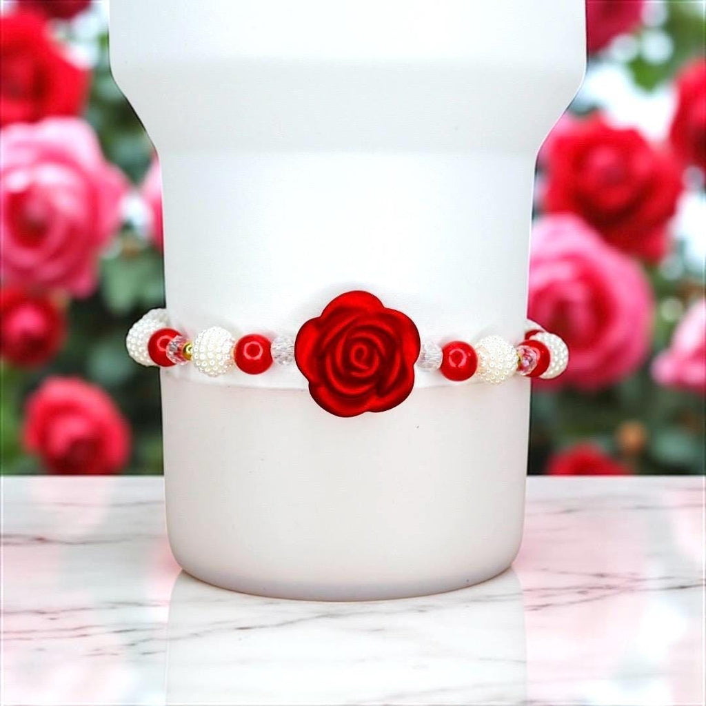 A white bracelet with red rose and bead accents. The bracelet features a large red rose in the center, surrounded by red and white beads. The bracelet is wrapped around a white object, with a blurred background of red roses.