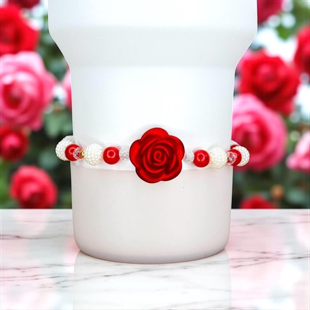 A white bracelet with red rose and bead accents. The bracelet features a large red rose in the center, surrounded by red and white beads. The bracelet is wrapped around a white object, with a blurred background of red roses.