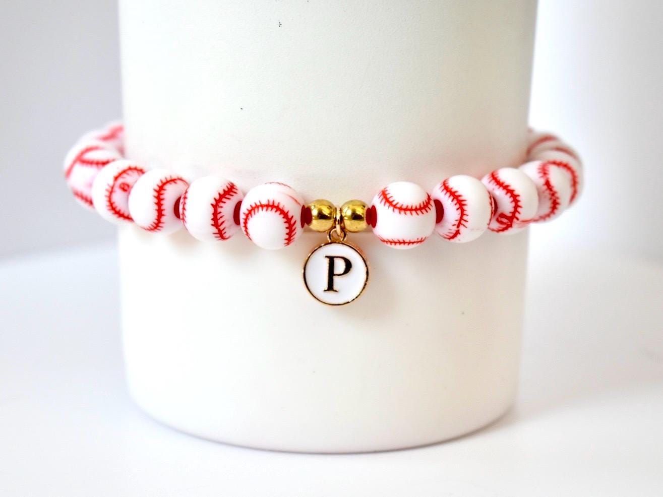 A baseball-themed bracelet featuring white beads with red stitching, strung together. A gold charm with the letter "P" hangs from the center. The bracelet is displayed on a white background.