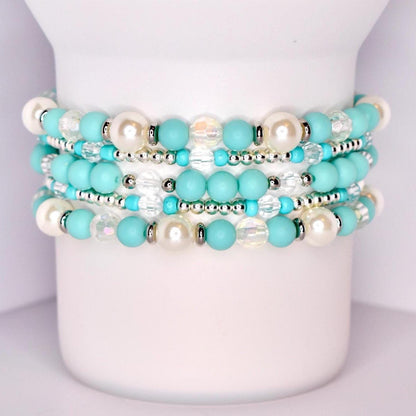 A set of five stacked tumbler bracelets displayed on a white tumbler, featuring a mix of turquoise matte beads, iridescent faceted beads, glossy pearl-like white beads, and shiny silver spacer beads for a vibrant, coastal-inspired design.
