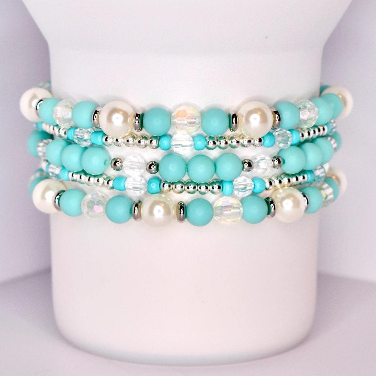 A set of five stacked tumbler bracelets displayed on a white tumbler, featuring a mix of turquoise matte beads, iridescent faceted beads, glossy pearl-like white beads, and shiny silver spacer beads for a vibrant, coastal-inspired design.